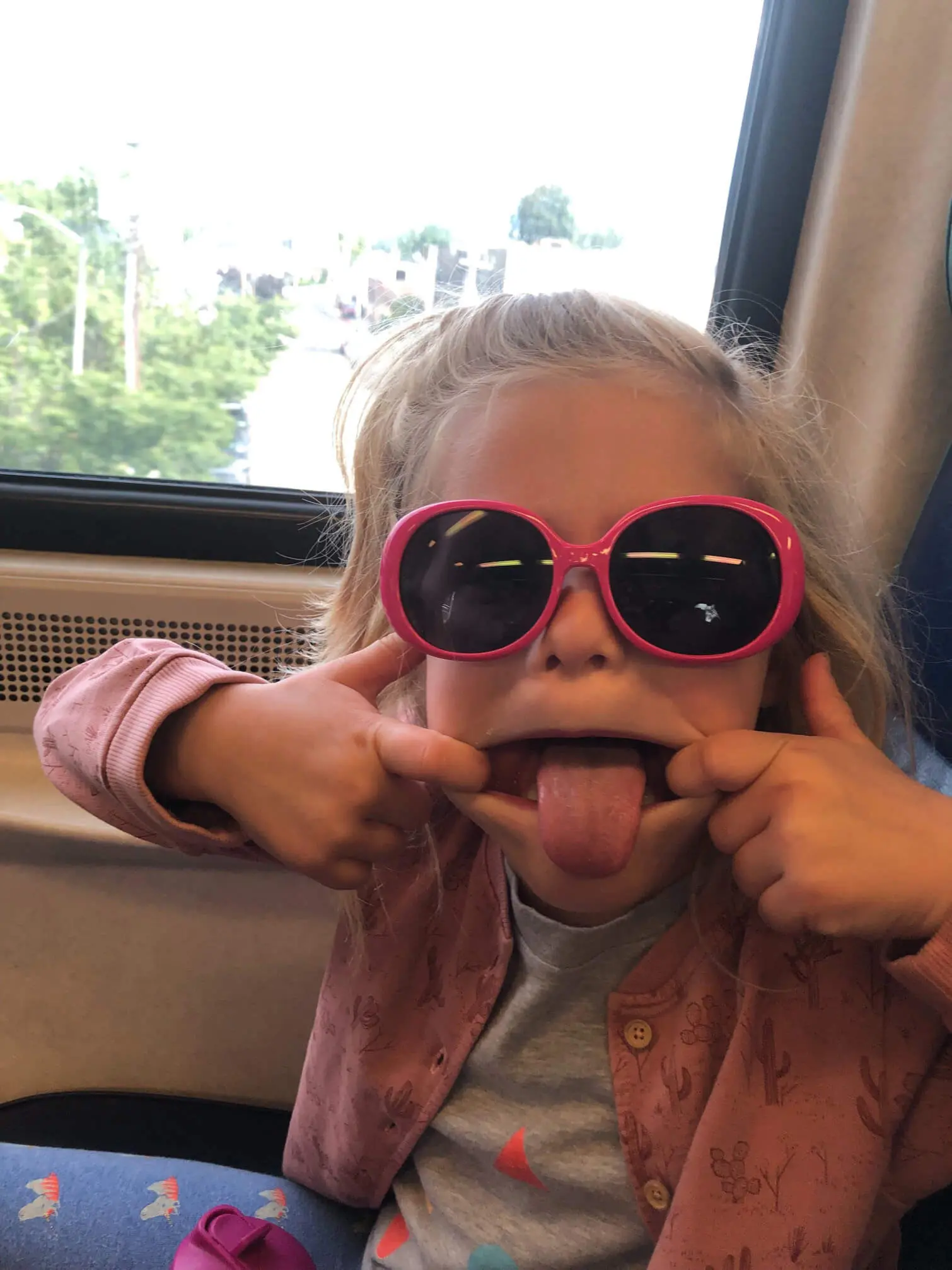 Young girl on a train, wearing sunglasses and making a silly face.